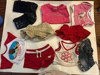 Nine pieces of Build-A-Bear clothing including shorts, skirts, tops, and denim jacket arranged on a white surface.