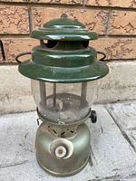 Side view of Coleman lantern showing green top vented cap and clear glass globe over the mantle inside. The silver fuel tank base shows wear and has a black adjustment knob.