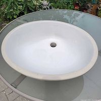 White ceramic oval undermount sink shown on glass table outside; central drain hole visible.