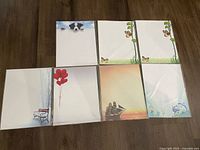 Seven unopened packages and various sheets of decorative 8.5x11 paper with different border designs and illustrations.
