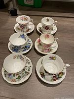 Top angle showing all six Royal Albert bone china teacups placed on matching saucers, each featuring different floral and fruit patterns in vibrant colors with gold trim.