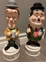 Front view of the two porcelain decanters representing Laurel and Hardy, showing facial features, clothing colors, and musical base with inscription.