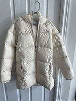 Front view of the lightly insulated chalk-colored puffer jacket with hood and zipper closure hanging on a hanger.