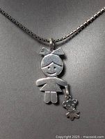 Close-up photo of sterling silver pendant featuring a girl figure with bow in hair and dangling teddy bear charm, on necklace chain.