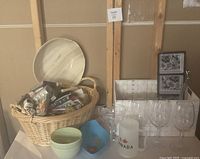 Photo showing large wicker basket with napkins, plates, plastic goblets, and other glassware displayed on a table.