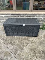 Front view of the black plastic outdoor storage deck box with a woven rattan texture, closed lid, and locking clasp on the lid.