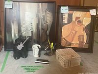 Two framed music-themed prints alongside music-related decor items: a black guitar sculpture, a white microphone mug, pens in a holder, and a rectangular box with printed sheet music pattern.