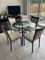 Glass top round dining table with black metal base and four beige upholstered chairs around it in dining room.