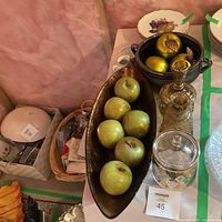 Photo of home decor set with artificial green apples in wooden tray, black ceramic bowl with golden decorative fruits, glass decanter, glass jar with floral design, stack of decorative plates, clear glass serving tray.