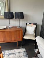 Photo showing two black lamps on a wooden cabinet next to a beige chair with a white pillow.