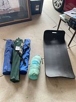 Top down view of five camping items laid out on garage floor: three wrapped camping chairs in blue and green bags, one black foam sleeping pad, one rolled sleeping pad in a mint green bag.