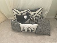 Seven decorative pillows arranged on the floor with white curtain backdrop, showing various patterns and colors in black, white, gray, and silver.