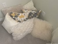 Seven decorative throw pillows in assorted sizes and colors including cream, white, yellow, and grey arranged in corner on floor