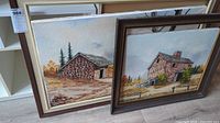 Two framed oil paintings leaning against wall on floor, showing rustic stone houses in natural landscapes