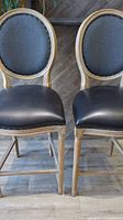 Front view showing pair of bar stools with oval backs upholstered in dark fabric and black padded seats with nailhead trim.