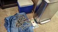 Blue electric blanket with controller, blue cooler, and carbon monoxide alarm on floor next to stainless steel bin