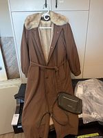 Full view of brown 3/4 length coat with fur lining and collar, hanging on hanger, with belt tied. Purse placed on table underneath coat.