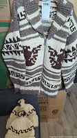 Full view of the handmade Cowichan sweater hanging with visible traditional designs and the two toques beneath it on a grey surface.