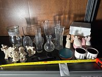 Photo showing assortment of ceramic figurines including nativity scene, glass vases of various shapes and sizes, bunny salt and pepper shakers, pastel coasters, and small ceramic cups with tree designs on shelf.