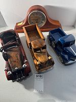 Three vintage model cars displayed in front of a wooden mantel clock on white surface.