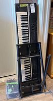 Rhodes keyboard leaning against a wall next to a black foldable keyboard stand and a plastic container with cables.