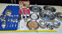 Various silver-plated cutlery, serving ladle, decorative trays, small bowls, salt and pepper shakers, and boxed fork and knife set displayed on blue cloth with measuring tape for scale.