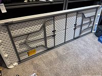 Underside of the folding table showing grey metal folding legs and frame attached to white plastic top.