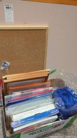 Box filled with office supplies including binders, folders, accordion files, plastic document holders, corkboard leaning behind the box, and a bundle of blue straps or cords.
