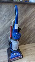 Side view of blue Hoover upright vacuum cleaner with attached hose and clear dust container.