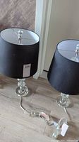 Two table lamps with glass bases and black shades on the floor next to a wall. Show their overall size, shape and cords.