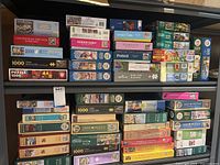 Photo showing multiple stacks of puzzle boxes arranged on shelves, brand names visible include Studio Puzzle, Piatnik, Ravensburger, Cobble Hill, and Ceaco. Puzzle counts range from 500 to 1500 pieces.