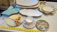 Top view of various porcelain and ceramic serveware items including plates with scenic designs and gold-trimmed dishes.