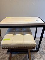 View of the small rectangular table with wooden top and black metal frame showing discoloration on top alongside the cream tufted stool beneath it.