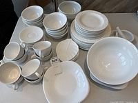 Photo of overall lot showing stacked white china cups, bowls, plates, a mortar and pestle, and a leaf-shaped serving dish on a table.