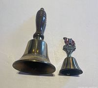 Two brass bells placed side by side on a white surface, showing the taller English bell with wooden handle and smaller Jerusalem bell with colorful emblem on top.