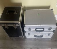 Two silver aluminum cases and one black heavy duty case side by side on floor.