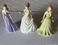 Front view of three Royal Doulton ladies figurines in purple, white, and green dresses on a white surface
