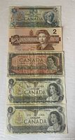 Five Canadian banknotes laid out showing front sides, denominations 1, 2, and 5 dollars, varying years including 1973, 1954, 1986, and 1979. Condition shows handling wear and creases.