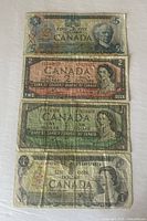 Four Canadian bills stacked showing front design: 1973 $1, 1954 $2, 1954 $1, 1979 $5 bill visible with wear and folding.