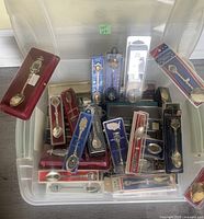 Plastic bin filled with multiple collectible souvenir spoons mostly in original packaging with varying designs visible.
