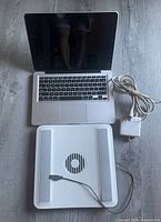 Top view of MacBook Pro laptop with black screen, keyboard, white fan cooling pad with rubber grips and USB cable, and white Apple charger