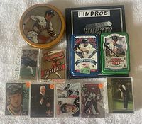 Overview photo of various sports cards displayed on a white towel including Roger Clemens cards in case, 1992 Bowman sealed pack, Lindros hockey tin and cards, Patrick Roy hockey tin and cards, Tiger Woods card in case, and other cards.