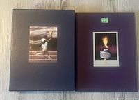 Two hardcover Olympic books, one black with torchbearer image and one dark purple with Olympic flame image on covers, resting side by side.