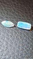 Pair of two natural Ethiopian opal gemstones, one cushion cut and the other marquis cut, showing multicolored flashes on a textured black surface.
