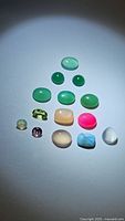Top-down photo of 15 individual polished natural gemstones arranged in a triangular pattern on a light surface under strong lighting, showing variety of green, pink, blue, beige and white colors, including cabochons and faceted stones.