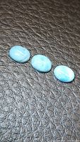 Three natural oval cabochon cut blue Larimar stones arranged in a row on a textured dark background, showing their smooth polished surfaces and varying light blue hues with white patterns.