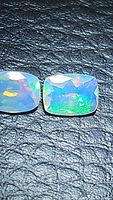 Close-up photo of two rectangular cushion-shaped faceted Ethiopian opal gemstones showing their multicolored flash on a black textured background.