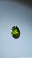 Oval cabochon cut apple green peridot gemstone shown on plain background. Visible internal clarity and surface reflection consistent with natural stone characteristics.
