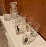 Angle showing the full vintage barware set arranged on white surface, including the cut glass decanter with stopper, two cut glass tumblers, two etched goblets, and four illustrated novelty highball glasses featuring British soldier motifs.