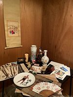 Photo showing assorted Japanese/Asian decorative items including porcelain vases, collectible geisha doll, decorative fans and plates set against a wood panel background.
